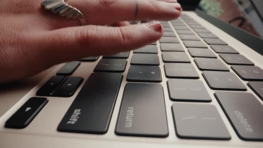 A close-up, low-angle view of a woman's hands with red nail polish and rings typing on a laptop keyboard
