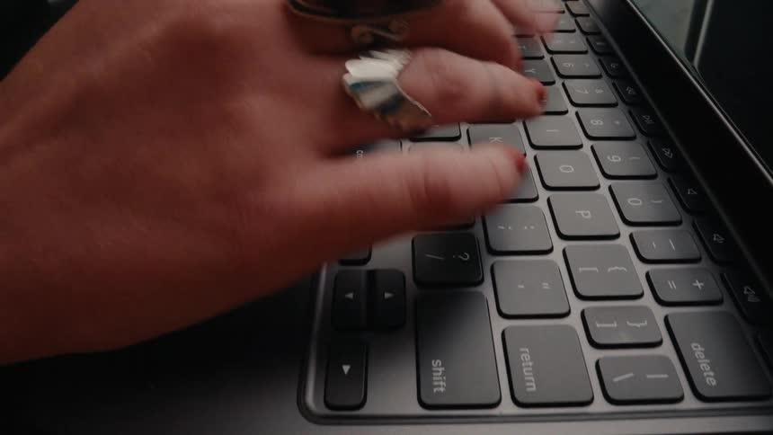 A close-up shot of a woman with red nail polish and ornate rings typing quickly on a tablet keyboard before closing the lid