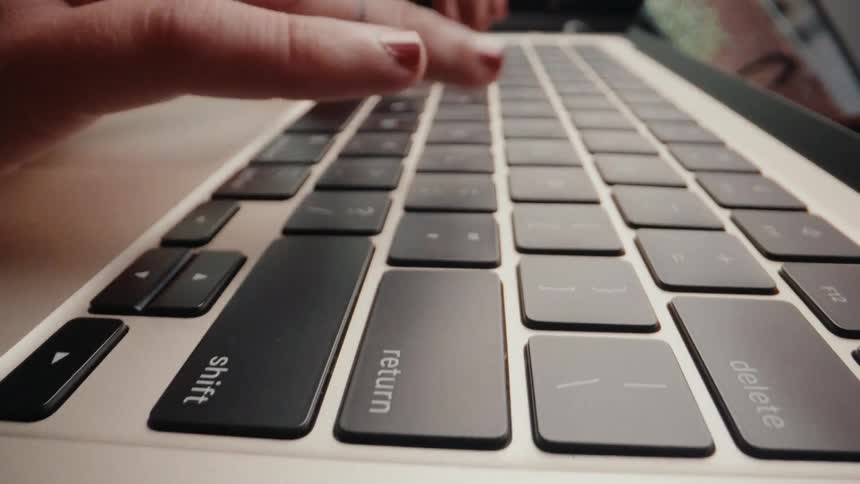A dramatic low-angle macro shot shows a person's hands with red nail polish and rings typing on a laptop keyboard