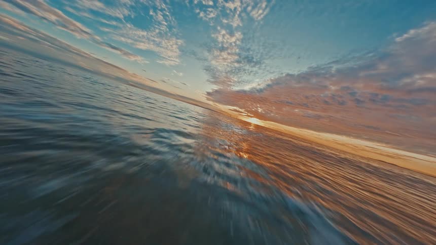 A breathtaking FPV drone flight skims low over the ocean surface towards a stunning sunset