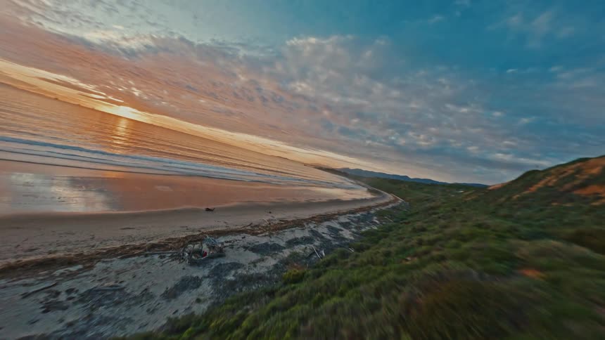 An exhilarating FPV drone flight along a lush coastal path reveals a breathtaking sunset over a tranquil ocean beach