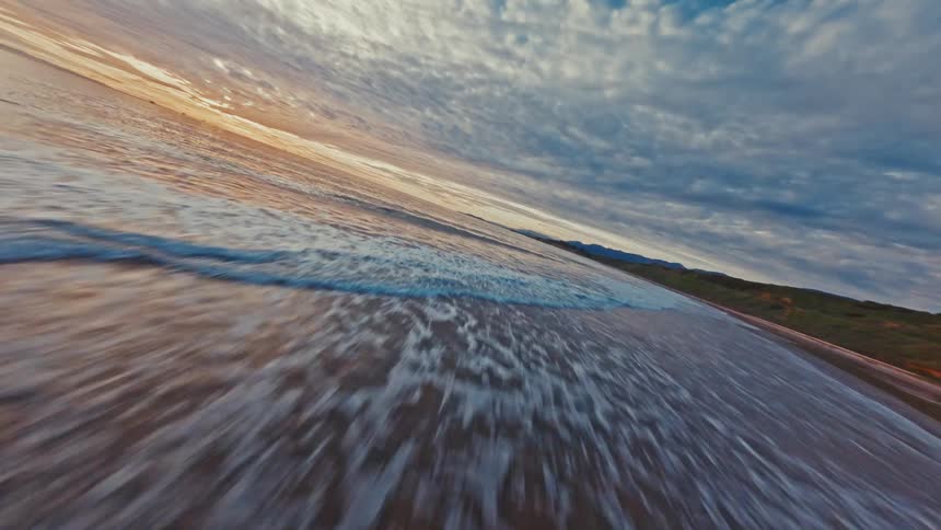 An exhilarating FPV drone flight skims low over the ocean waves and sandy shoreline under a dramatic, cloud-filled sunset sky