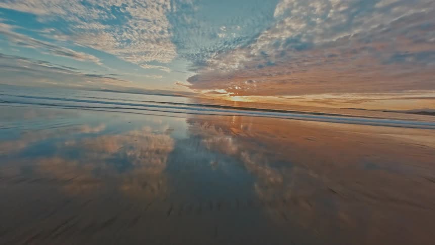 An exhilarating FPV drone flight swoops down from a coastal cliff, racing across a sandy beach and out over the ocean waves towards a stunning sunset