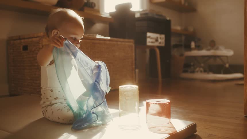 A cute baby sits on the floor in a warm, sunlit room and curiously plays with a sheer scarf in slow motion