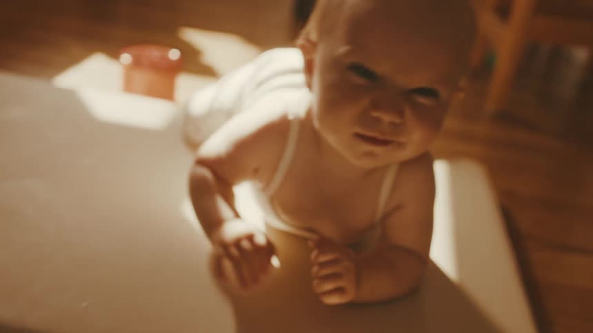 An adorable baby with big blue eyes lies on its tummy in a sunlit room, looking at the camera