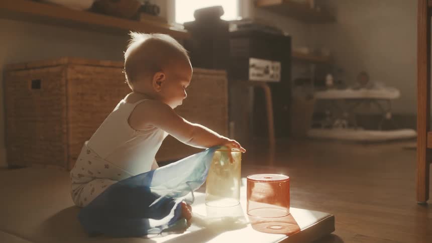A curious baby explores a colorful scarf and cups while sitting in a warm sunbeam on the floor