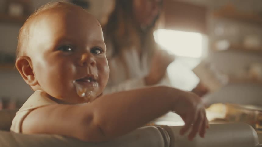 A cute baby with a messy face looks at the camera while being spoon-fed by its mother in a high chair