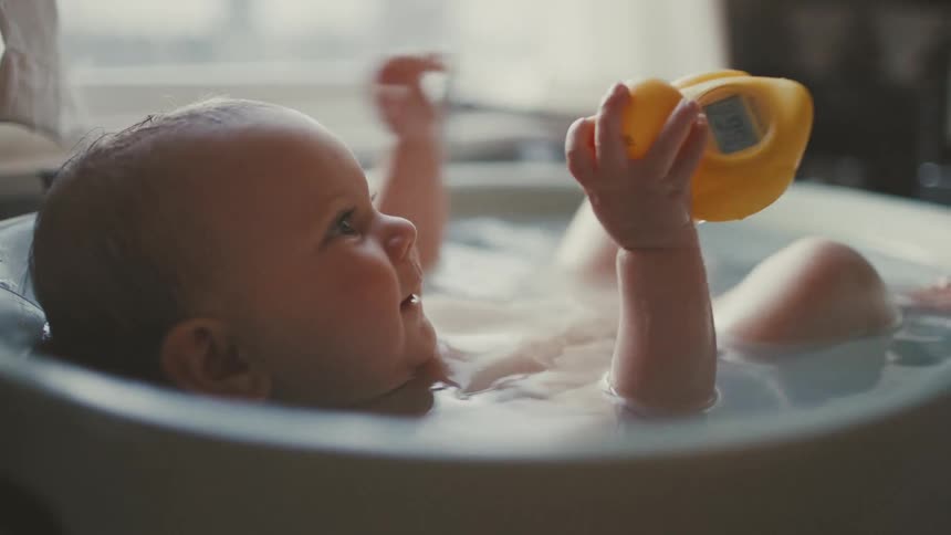 An adorable smiling baby lays on their back in a bath playing with a yellow rubber duck