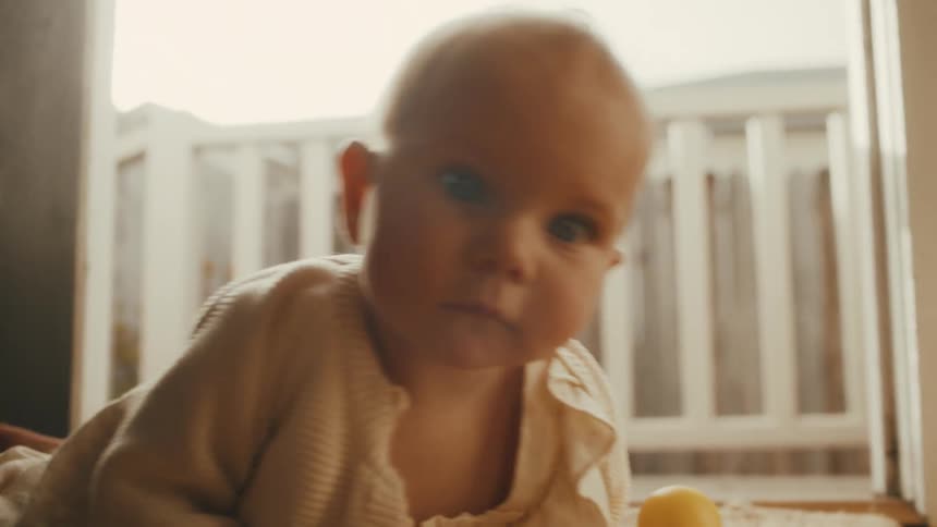 An adorable baby with curious blue eyes lies on a warm, sunlit rug and begins to crawl towards the camera with a yellow rubber ducky