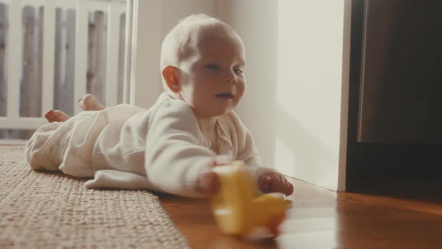 A cute baby lies on her tummy on a rug and hardwood floor, playing with a yellow rubber ducky in slow motion