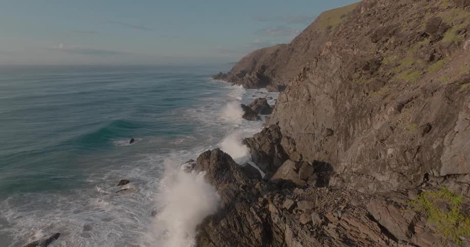 Powerful ocean waves crash in slow motion against a rugged, rocky cliffside at sunrise