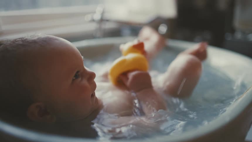 A cute smiling baby lays back in a small tub, splashing happily with their feet and holding a yellow rubber duck in slow motion