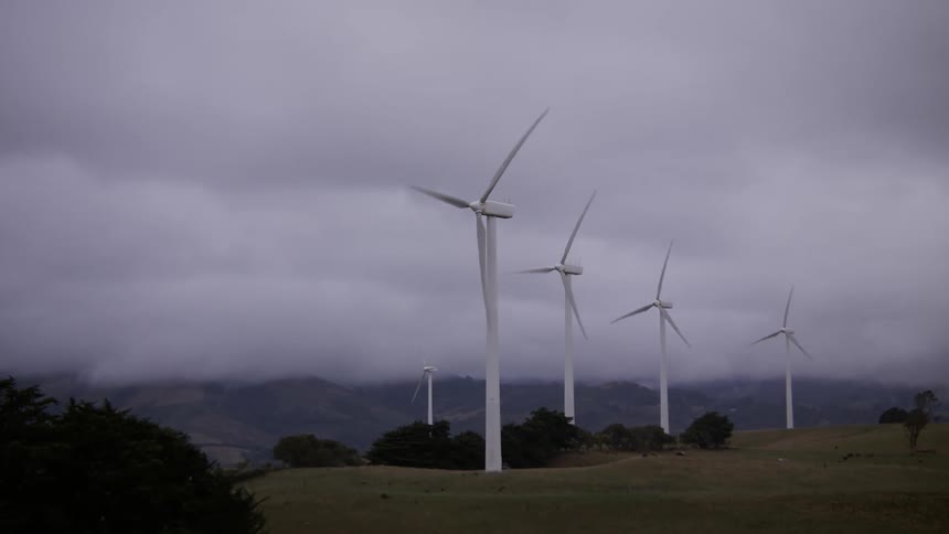 Wind turbines spin on a grassy hillside, generating renewable energy under a moody, overcast sky with mountains in the background