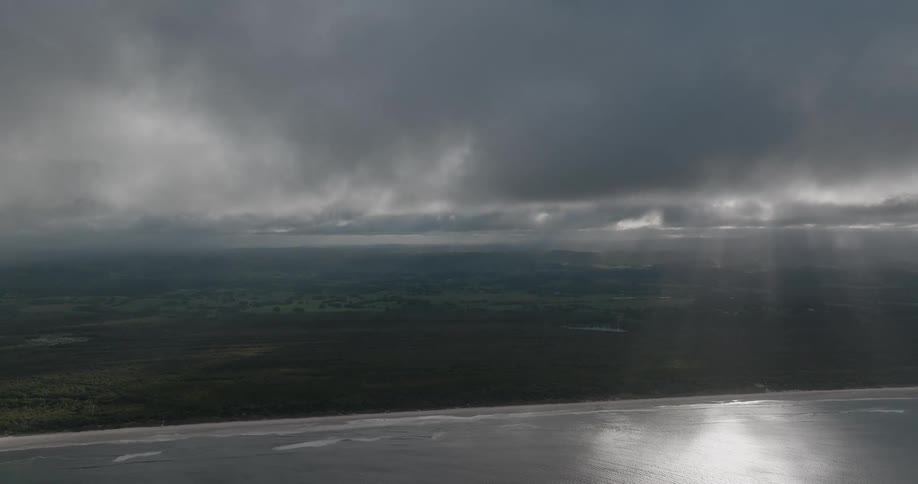 Sunbeams shine through stormy clouds over a wide ocean coastline and verdant landscape in a stunning aerial view
