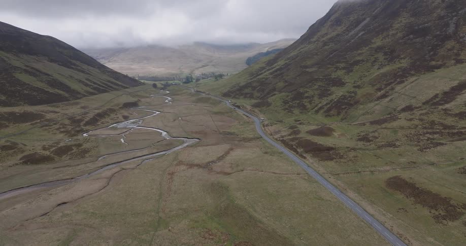 A static aerial view shows a winding river and a quiet road running through a vast, green valley in the Scottish Highlands on an overcast day