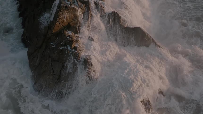A dramatic overhead slow-motion shot captures the raw power of ocean waves crashing against rugged coastal rocks
