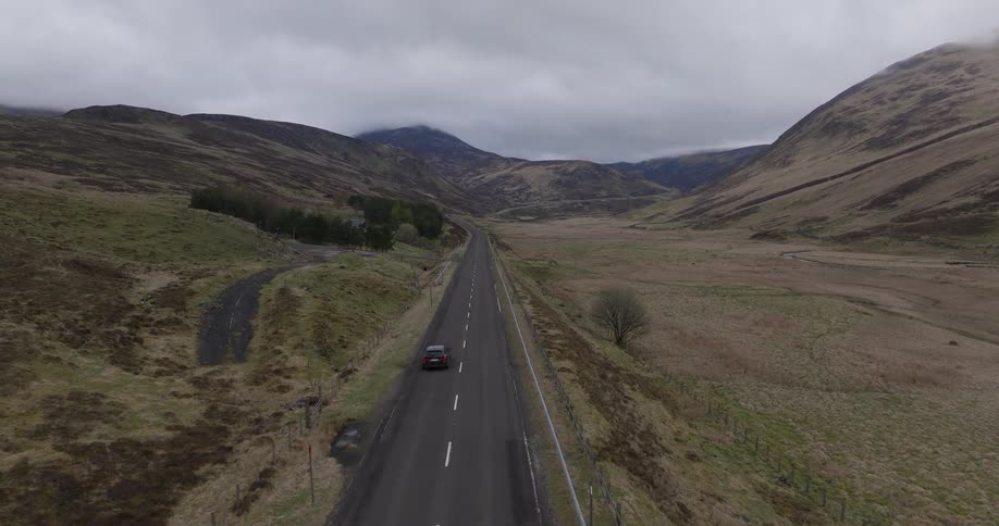 Aerial footage follows a car driving along a remote highway through a vast mountain valley under a moody, overcast sky