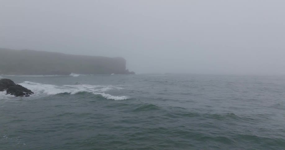 A slow motion view of the ocean with waves crashing against a rocky coastline on a very foggy day