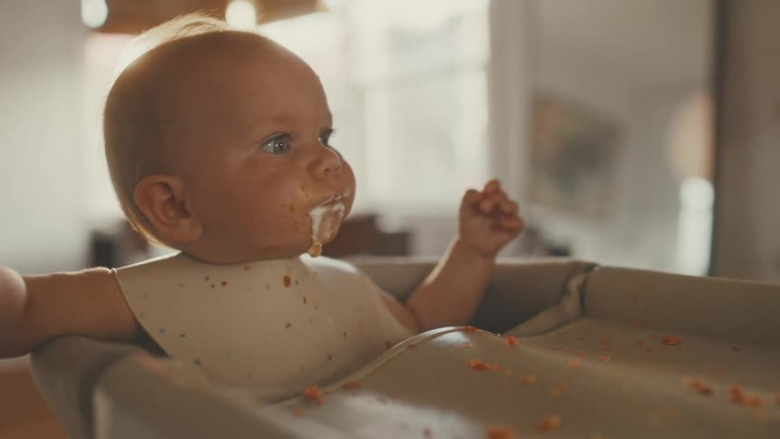 Slow motion close up of a cute messy baby in a high chair being spoon-fed by a parent in a sunlit room