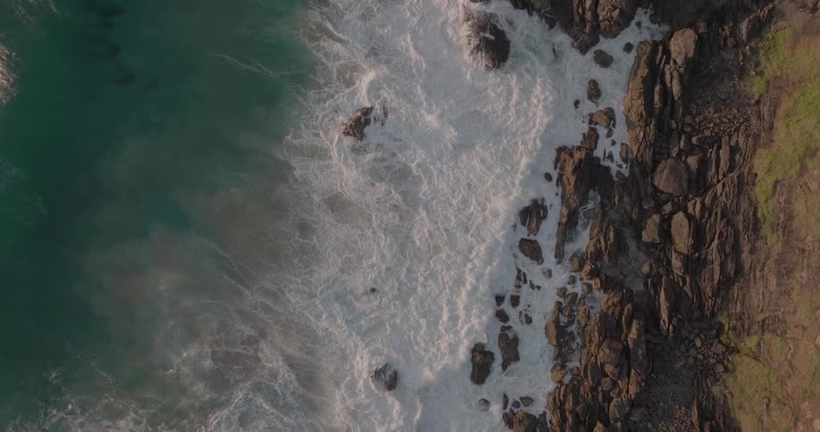 A powerful aerial top-down perspective shows turquoise ocean waves crashing in slow motion against a rugged, rocky coastline