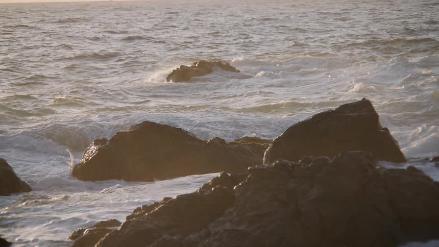 Slow motion footage shows powerful ocean waves crashing over dark, jagged rocks during a beautiful golden hour sunset