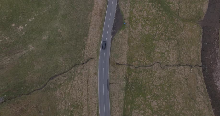 A top-down aerial view follows a black car driving along a paved road through a vast, green rural landscape