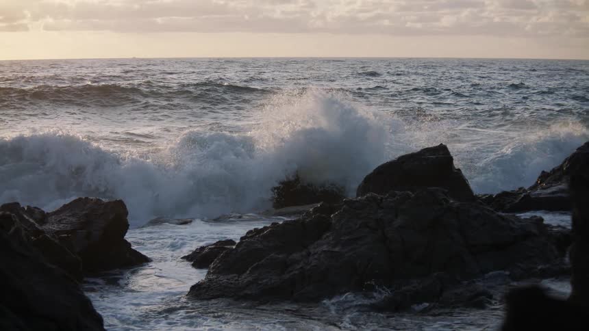 Slow motion view of a powerful ocean wave breaking over dark coastal rocks at sunset