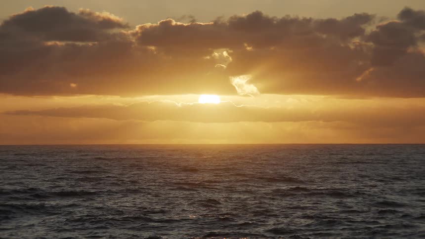 A tranquil and beautiful golden sunset over the open ocean as the sun peeks through the clouds