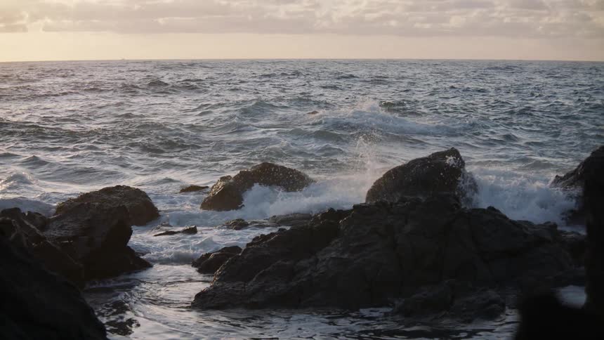 Dramatic slow-motion view of ocean waves crashing over a rocky shoreline at sunset