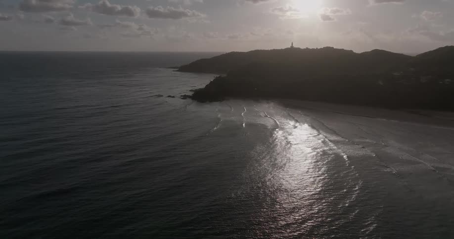 A serene aerial view shows the sun reflecting off the ocean as gentle waves lap against the shore of a silhouetted coastline at dusk