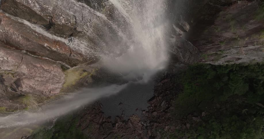 A breathtaking top-down aerial shot reveals a powerful waterfall cascading down a sheer rock cliff into a pool below
