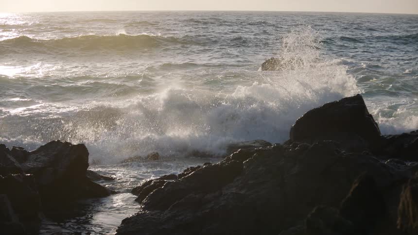 Golden hour sun illuminates the ocean as powerful waves crash against a rocky shoreline in slow motion