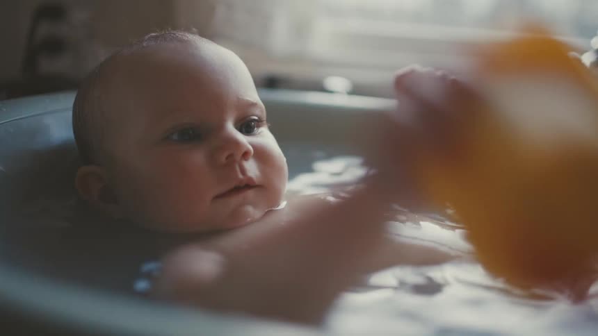 An adorable baby boy smiles while lying in a small tub playing with a yellow rubber duck in slow motion