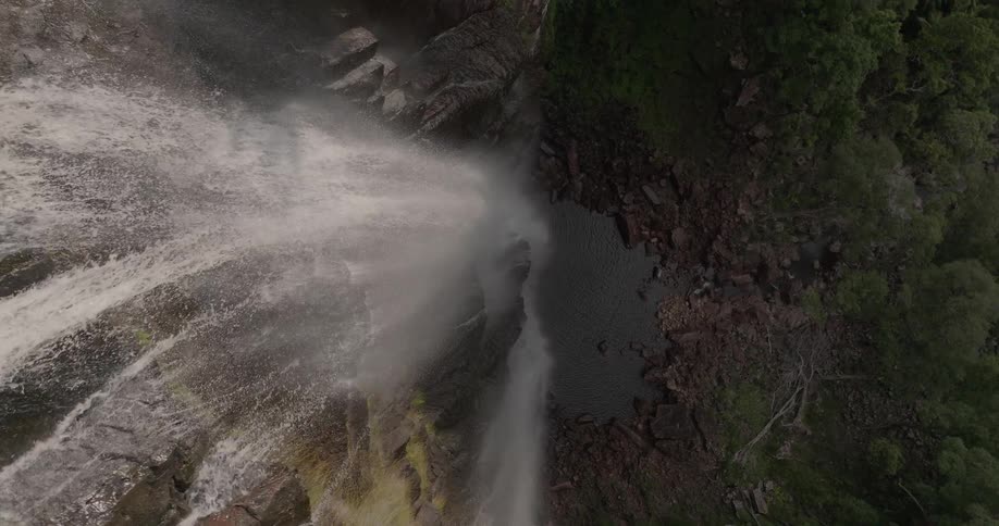 A dramatic top-down aerial shot slowly rises above a powerful waterfall crashing onto the rocks below
