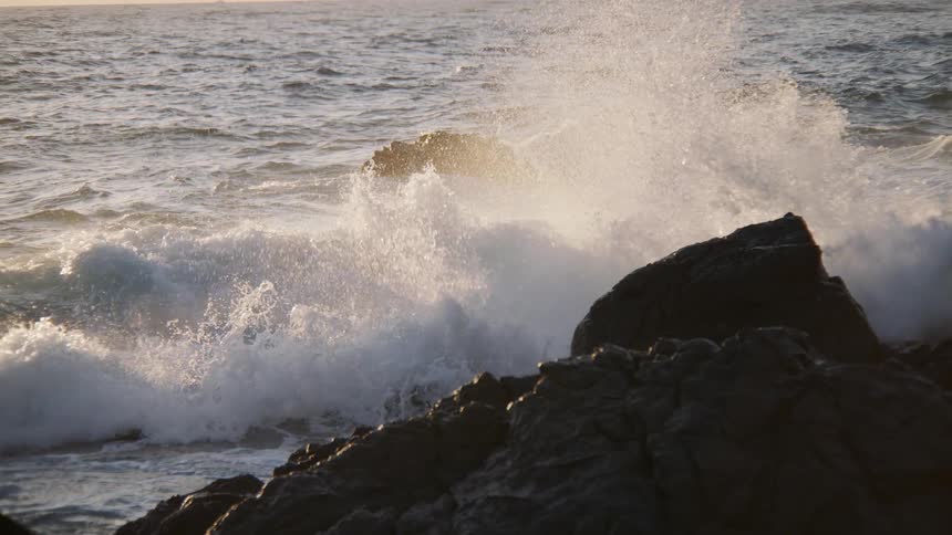 A powerful, sunlit ocean wave crashes against dark coastal rocks in dramatic slow motion