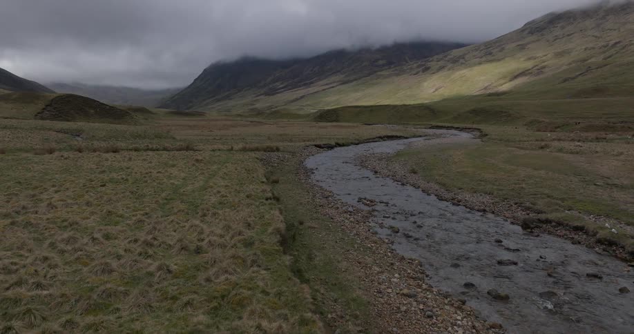 An aerial view following a meandering river through a vast, moody valley in the Scottish Highlands under a cloudy sky
