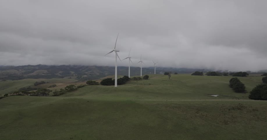 Wind turbines turn slowly on a vast green landscape under a moody, overcast sky
