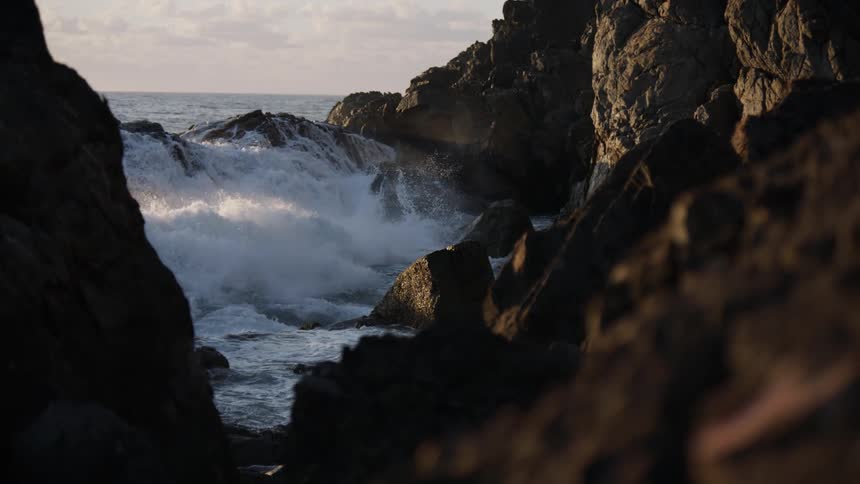 A powerful ocean wave crashes against a rocky coastline in dramatic slow motion during a beautiful sunset