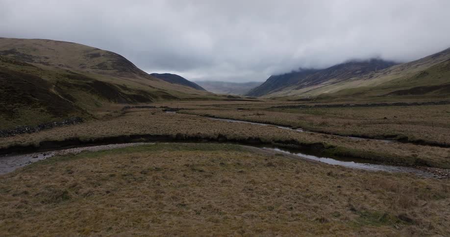 An aerial view flying over a winding river through a vast, remote valley in the Scottish Highlands on a cloudy day