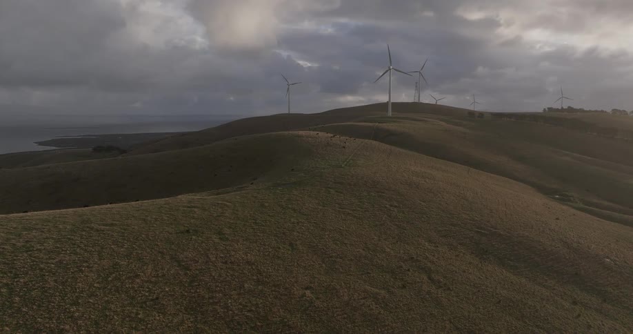Wind turbines stand majestically on rolling coastal hills under a dramatic and cloudy sky as the camera slowly pushes forward