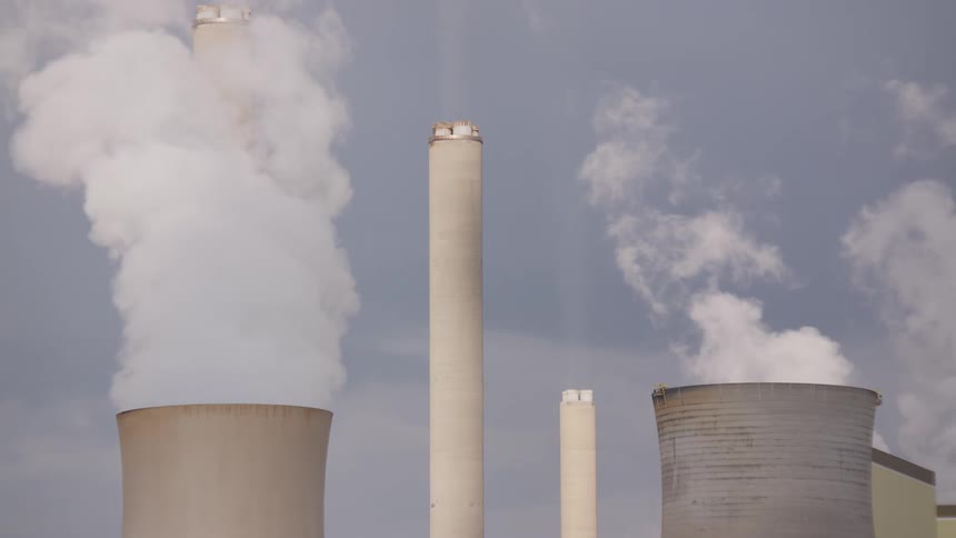 Steam and smoke rise from the cooling towers and chimneys of a power plant against a cloudy sky