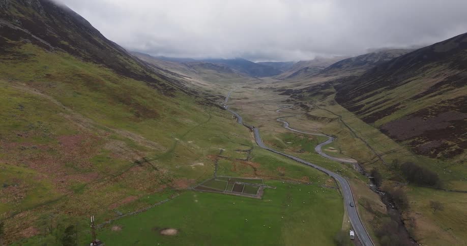 Cinematic aerial footage reveals a vast, green mountain valley with a serpentine road and river under a cloudy sky