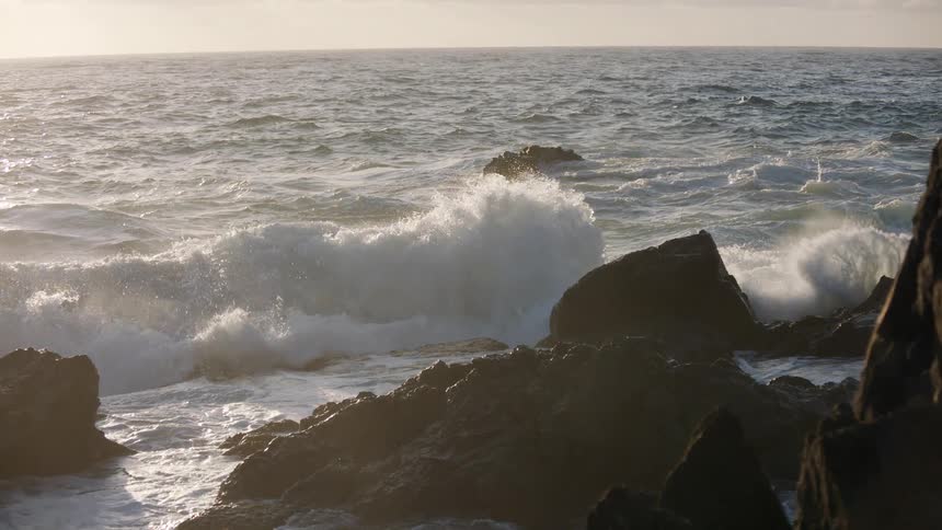 A dramatic slow-motion shot captures the moment a large ocean wave crashes against a rocky shore, backlit by the setting sun