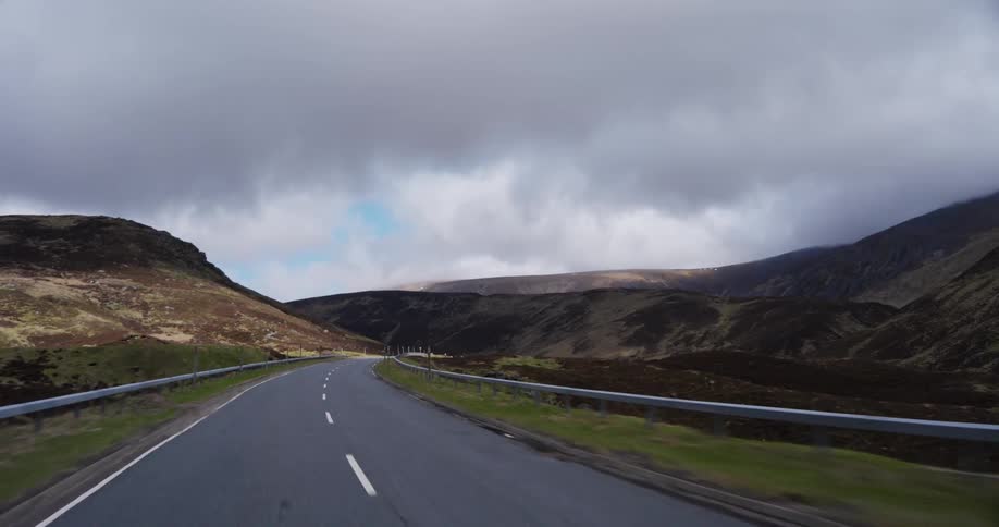A point-of-view shot driving along a scenic road that winds through the rugged Scottish Highlands on an overcast day