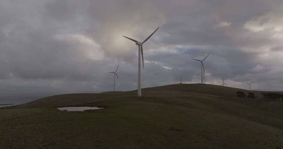 A dramatic slow push-in shot shows wind turbines spinning on a coastal hillside under a moody and overcast sky