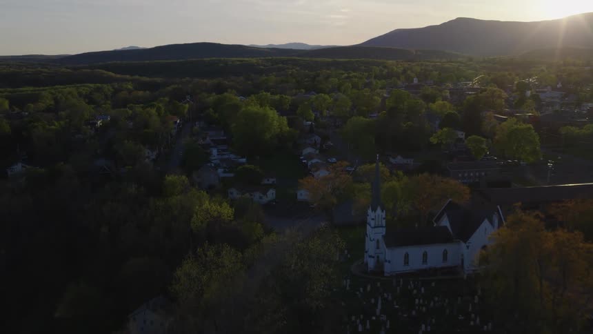 Aerial view of a quaint small town featuring a white church and cemetery during a beautiful sunset over the mountains