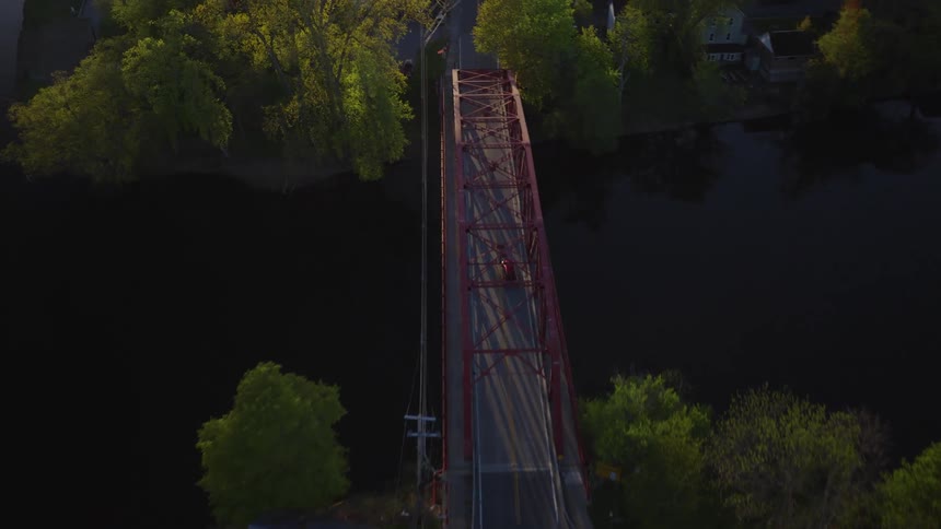 Aerial top-down view of cars driving across a red truss bridge over a dark river during a peaceful golden hour sunset