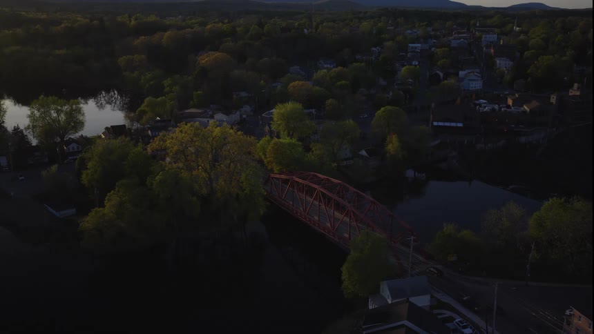 A tranquil aerial view shows a charming small town nestled among lush green trees, with a red bridge crossing a river during a golden sunset