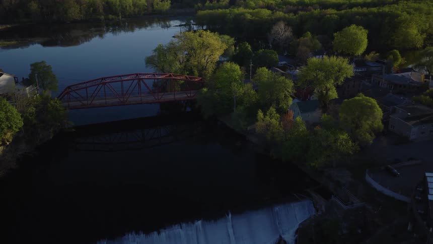 A scenic aerial view pushes in on a waterfall and red bridge crossing a river next to a small town at sunrise