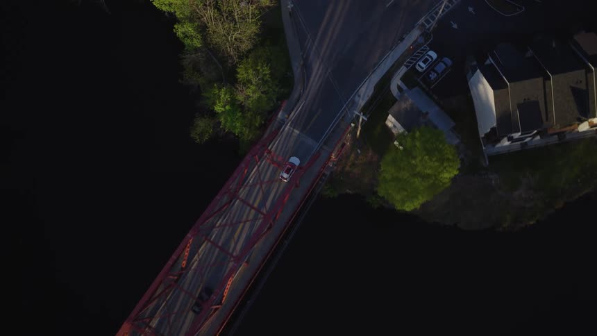 Aerial top-down view of a car driving across a red truss bridge over a river into a small town during golden hour